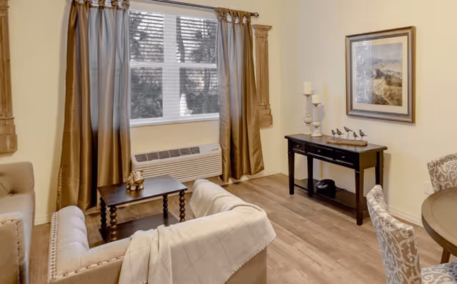 A cozy living room area with a beige sofa draped with a light-colored blanket, a small dark wooden coffee table, and a window with brown curtains. There is a black console table against the wall with decorative items and candles, a framed landscape painting above it, and part of a round dining table with patterned chairs visible on the right. The room has light-colored walls and wood flooring.