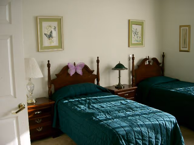 A bedroom with two twin beds featuring dark wooden headboards and green bedspreads. Each bed has a nightstand beside it, one with a white lamp and the other with a green lamp. There are three framed pictures hanging on the wall above the beds.