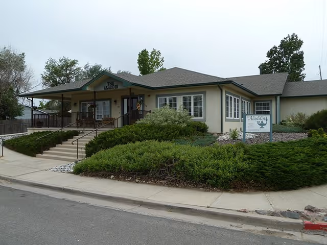 Front exterior of a single-story assisted living facility with a covered porch, ramp and steps, landscaped shrubs, and an 'Aladdin' sign.