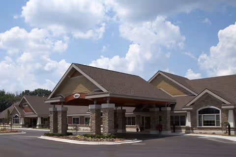 Exterior view of a single-story senior living facility building with a covered entrance supported by stone pillars, a brown roof, and beige siding under a partly cloudy sky.
