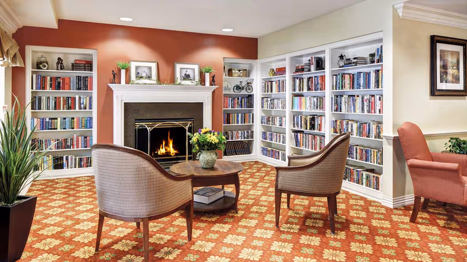A cozy library room with a lit fireplace in the center, flanked by white built-in bookshelves filled with books. Two cushioned armchairs face a small round wooden table with a flower vase on it. The room has a patterned carpet with floral designs and warm-colored walls.