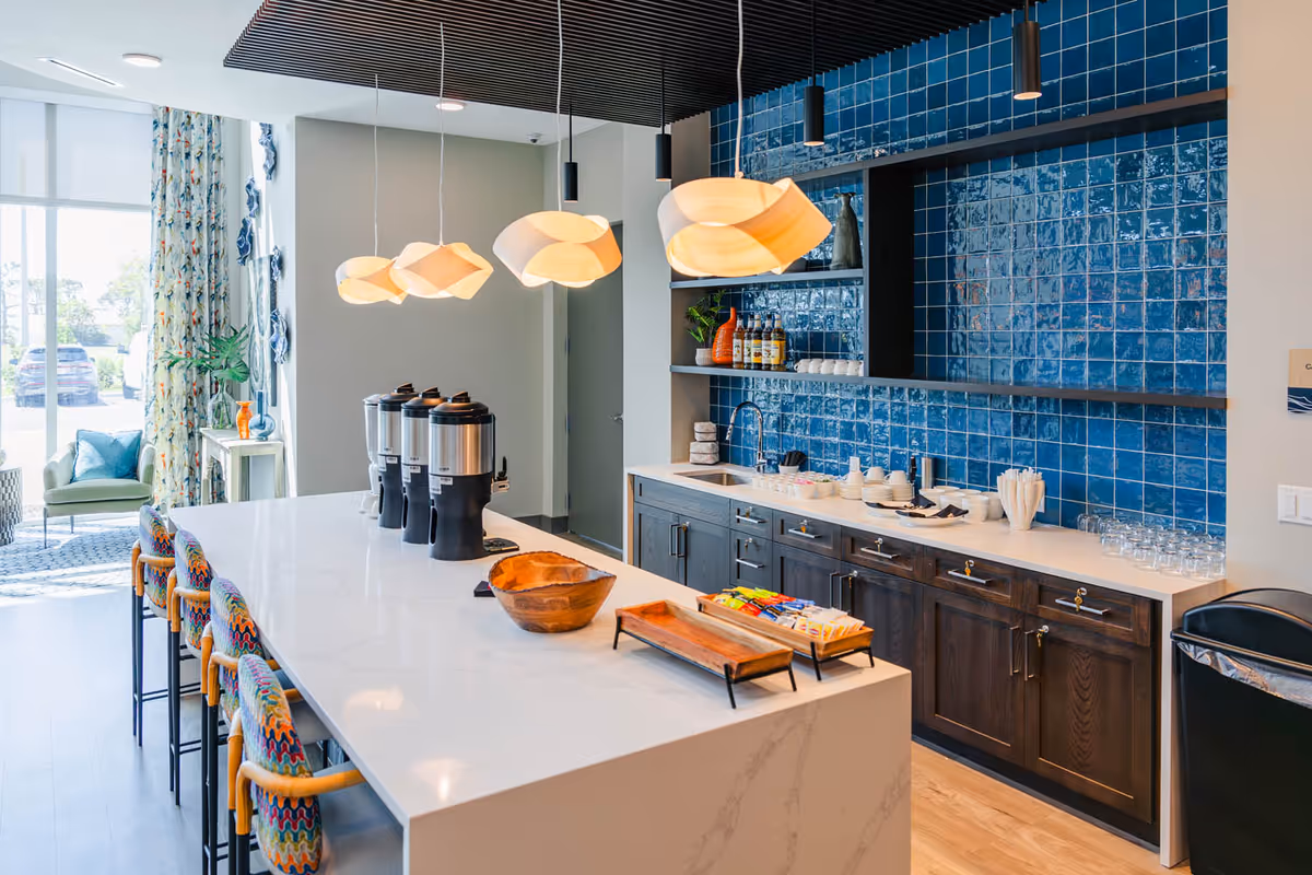 Modern kitchen area with a long white marble island countertop featuring colorful bar stools, three beverage dispensers, and wooden trays. The back wall has dark wood cabinets and shelves with blue tiled backsplash, decorated with bottles, cups, and plants. Pendant lights hang from the ceiling, and a large window with floral curtains lets in natural light.