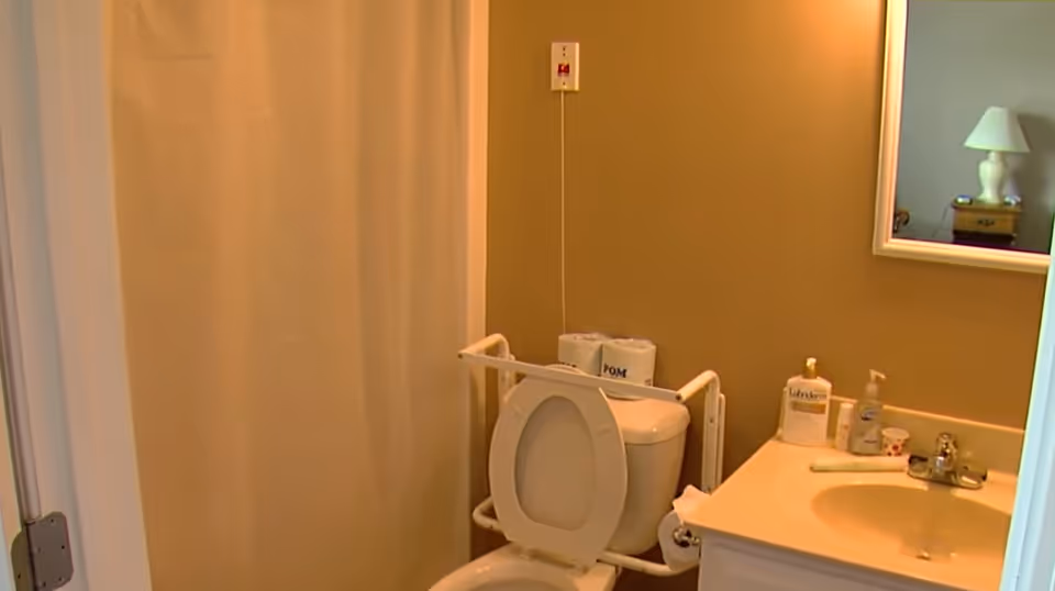 Small accessible bathroom with a toilet and safety rails, sink with toiletries and mirror, and a shower curtain.