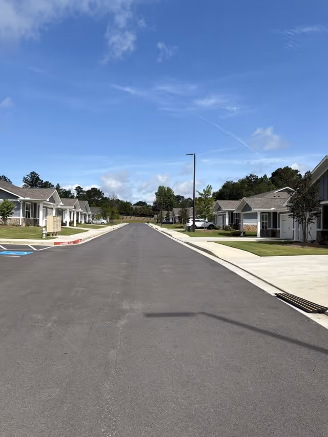 A paved road lined with single-story residential buildings on both sides under a blue sky with some clouds. The buildings have front porches, driveways, and small lawns with trees and shrubs.
