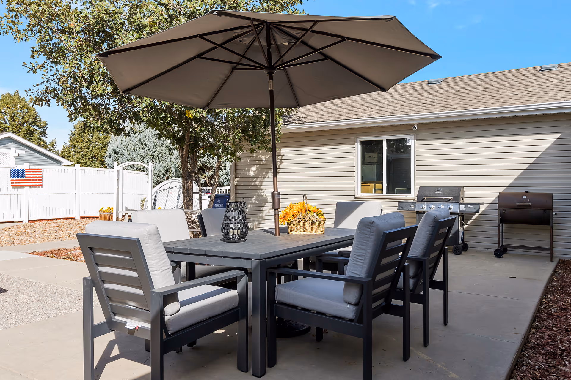 Outdoor patio area with a rectangular table surrounded by six cushioned chairs. A large umbrella is positioned in the center of the table providing shade. On the table, there is a decorative lantern and a basket with yellow flowers. In the background, there are two grills against the beige siding of a building, a white fence, and some trees.