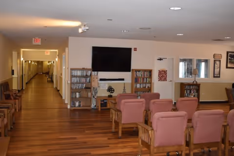 A common area in Orchards Assisted Living featuring several cushioned chairs arranged in rows facing a wall-mounted flat screen TV. There are wooden shelves with DVDs and books on either side of the TV. The room has wood flooring and beige walls with a hallway extending to the left with multiple doors and exit signs visible.