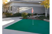 Outdoor putting green with a red flag in front of a building with stone and gray siding, surrounded by shrubs and trees with autumn leaves.