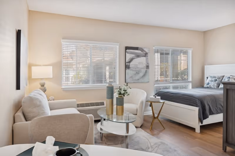A bright and cozy studio apartment featuring a white sofa, a white armchair, a glass coffee table with decorative vases, a small side table with books, and a white bed with gray bedding. Two windows with white blinds allow natural light to fill the room, which has light-colored walls and wooden flooring.