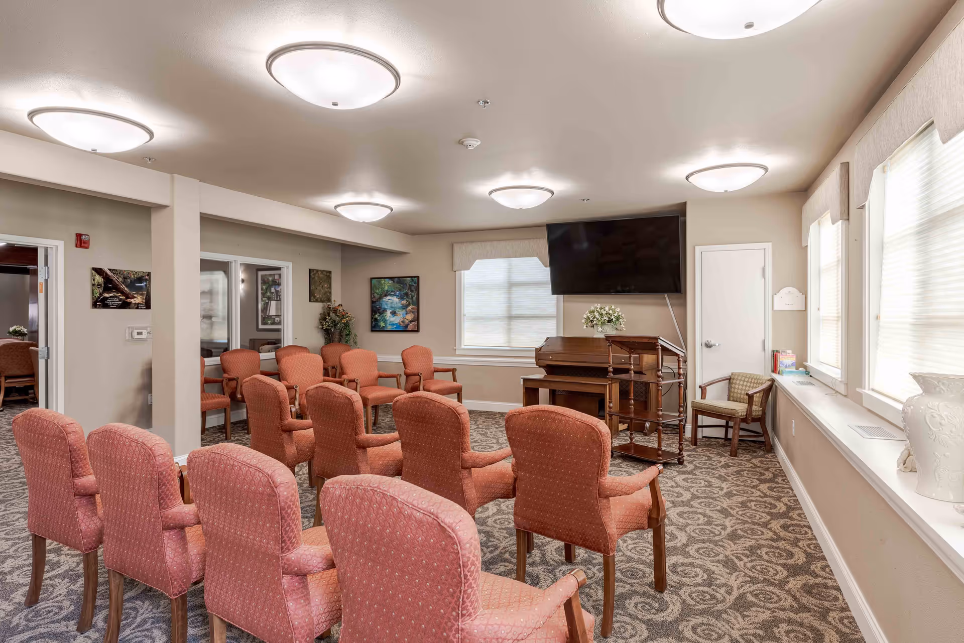 A well-lit common room with multiple upholstered chairs arranged in rows facing a piano and a large wall-mounted TV. The room has patterned carpet, beige walls, several windows with blinds, and ceiling lights. There are framed pictures on the walls and a small table with flowers near the piano.
