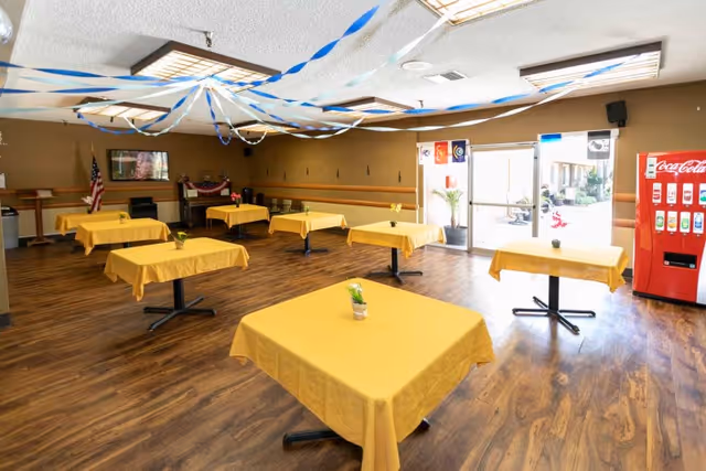 A spacious dining room with several square tables covered in yellow tablecloths, each with a small potted plant centerpiece. The room has wood flooring, an American flag, a piano, and blue and white streamers hanging from the ceiling. A red Coca-Cola vending machine is visible near the entrance door, which lets in natural light.