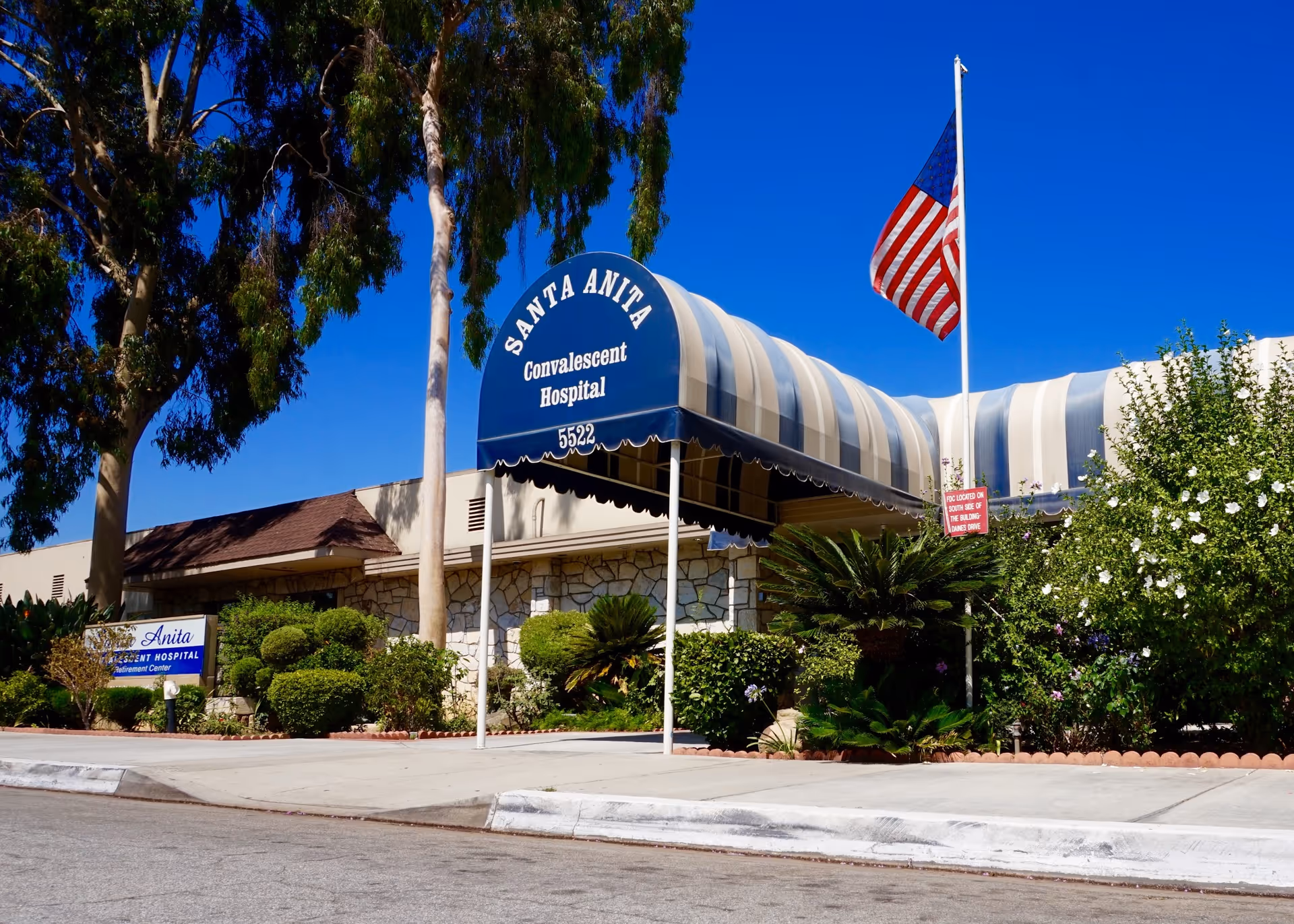Exterior view of Santa Anita Convalescent Hospital with a blue and white striped awning over the entrance, an American flag on a flagpole, and well-maintained bushes and trees under a clear blue sky.