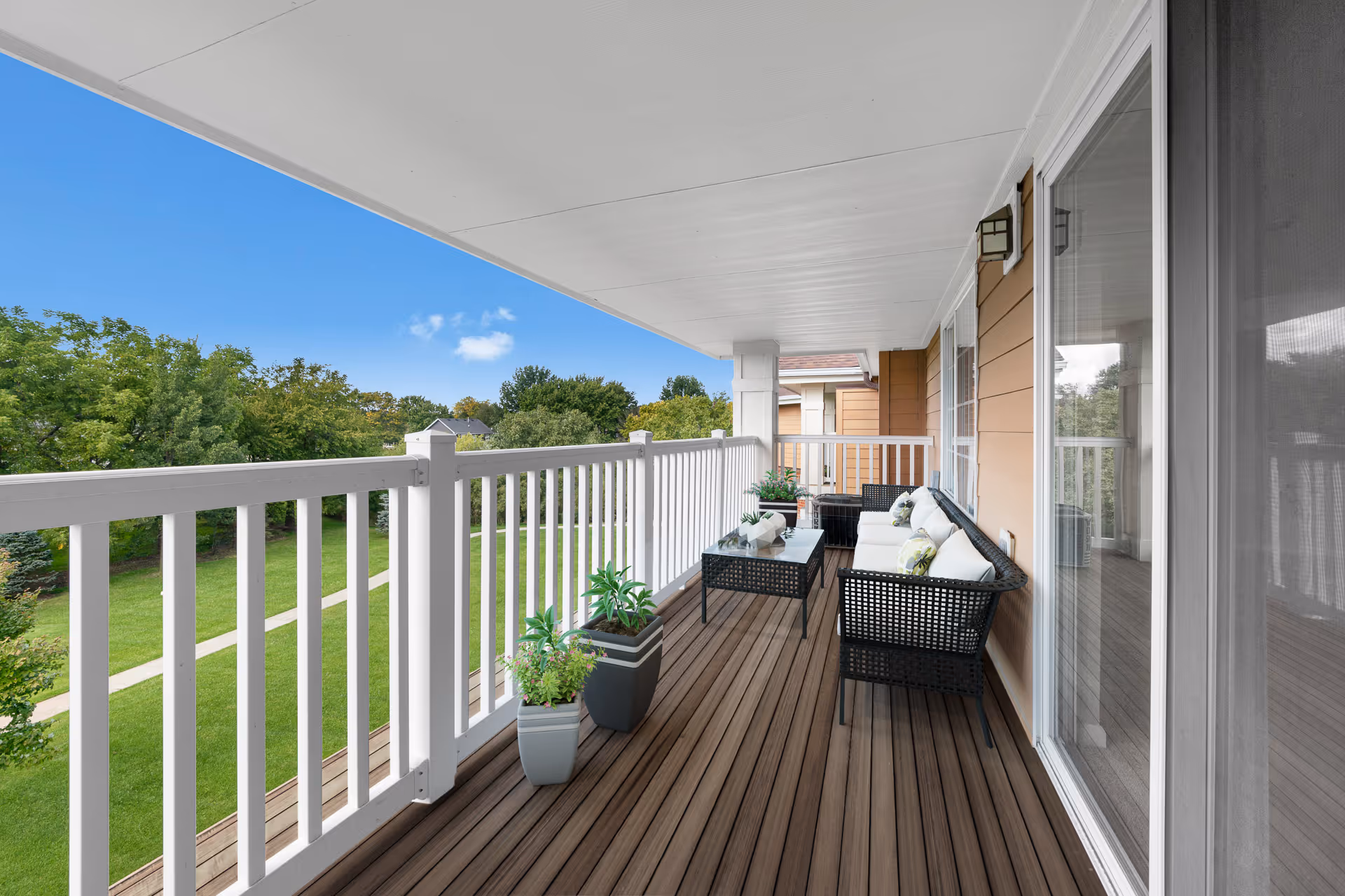 Covered balcony with wooden flooring, white railing, black wicker furniture with white cushions, glass-top coffee table, and potted plants overlooking a green lawn and trees under a clear blue sky.