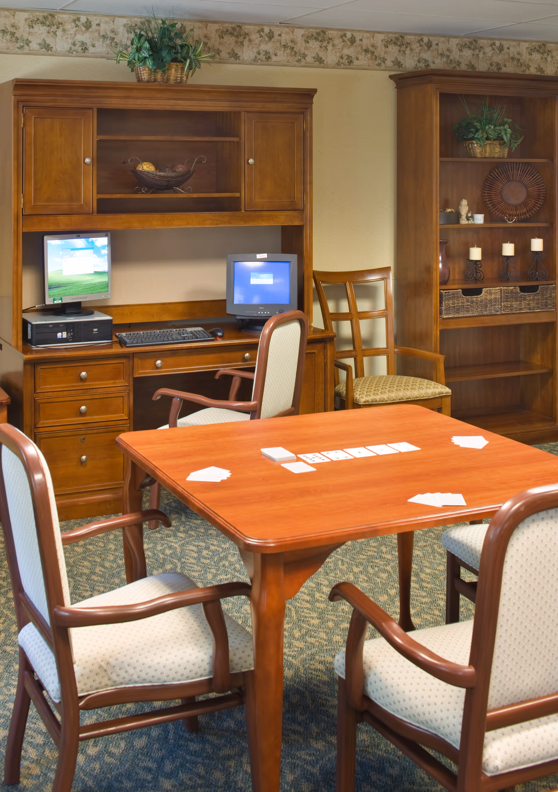 A cozy room with a wooden table and four cushioned chairs arranged around it. On the table, there are playing cards laid out. In the background, there is a wooden desk with two computer monitors and a keyboard, along with a wooden bookshelf decorated with plants, candles, and baskets. The room has a carpeted floor and wallpaper with a floral border near the ceiling.