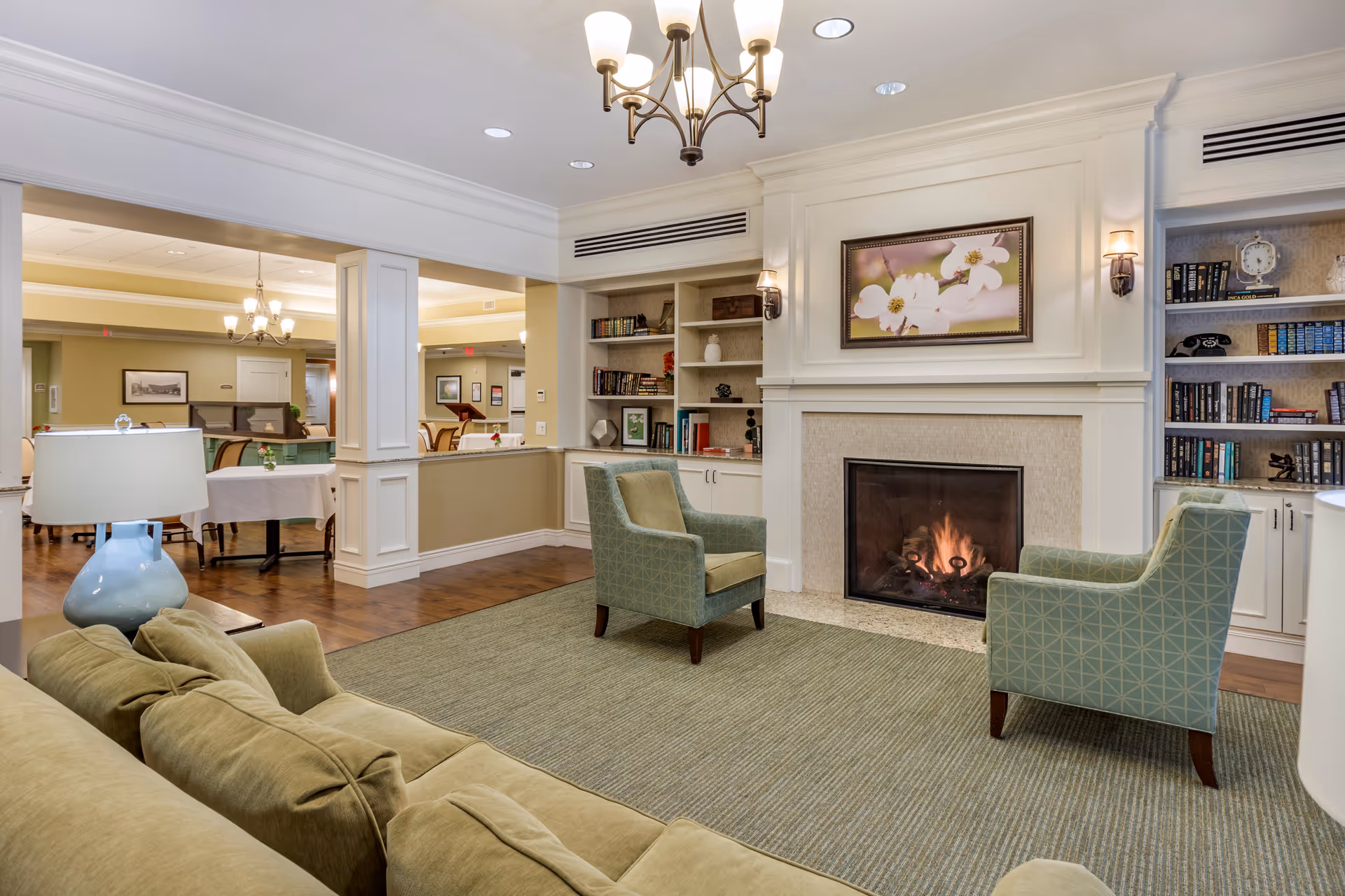 Common area lounge with a lit fireplace, two armchairs and a sofa, bookshelves, and a dining area visible in the background.
