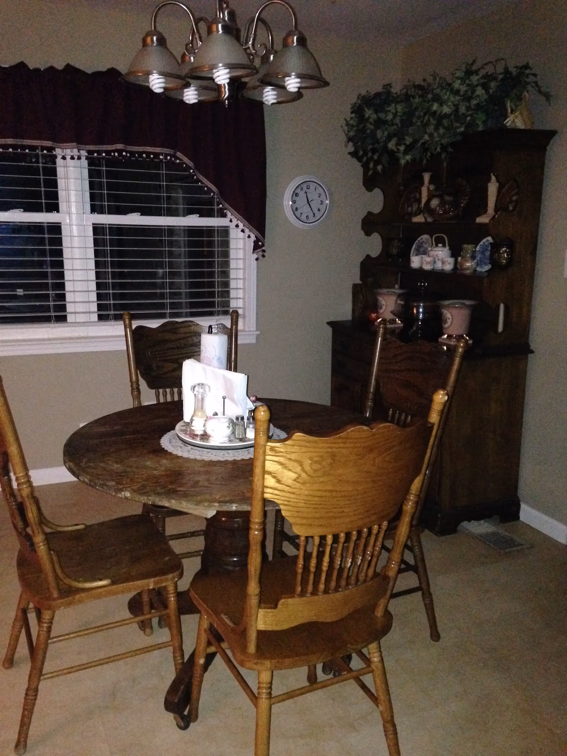 Wooden round dining table with four chairs under a chandelier next to a hutch and a window with blinds.