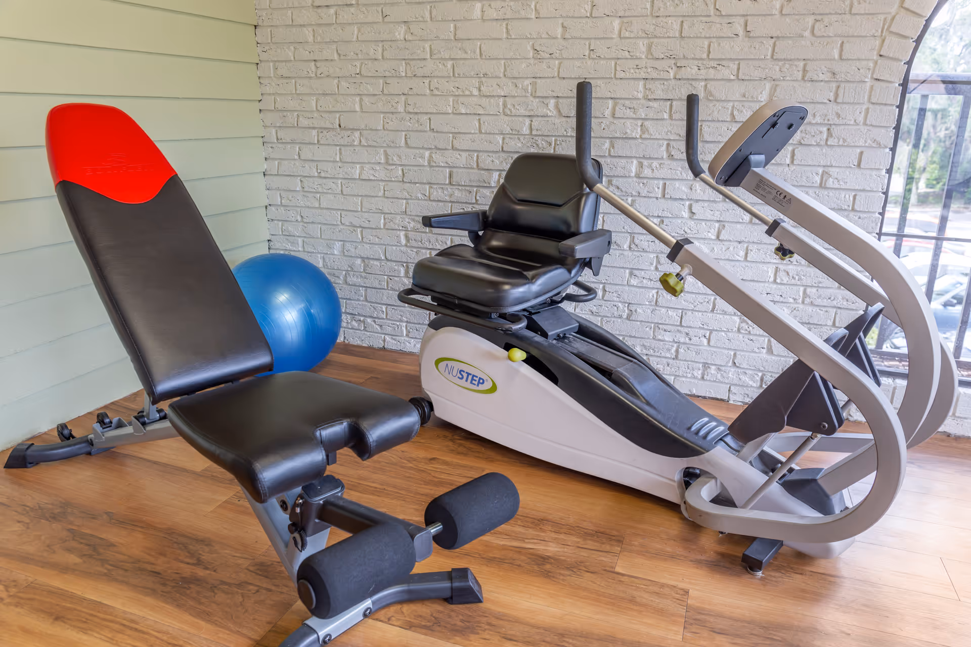 Small exercise room with an adjustable workout bench, a blue stability ball, and a NuStep recumbent cross-trainer against a white brick wall.