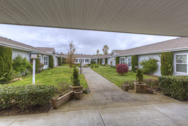 View of a senior living facility courtyard with a concrete walkway leading through a well-maintained lawn and landscaped bushes and trees, surrounded by single-story buildings with light-colored siding and multiple windows under a cloudy sky.