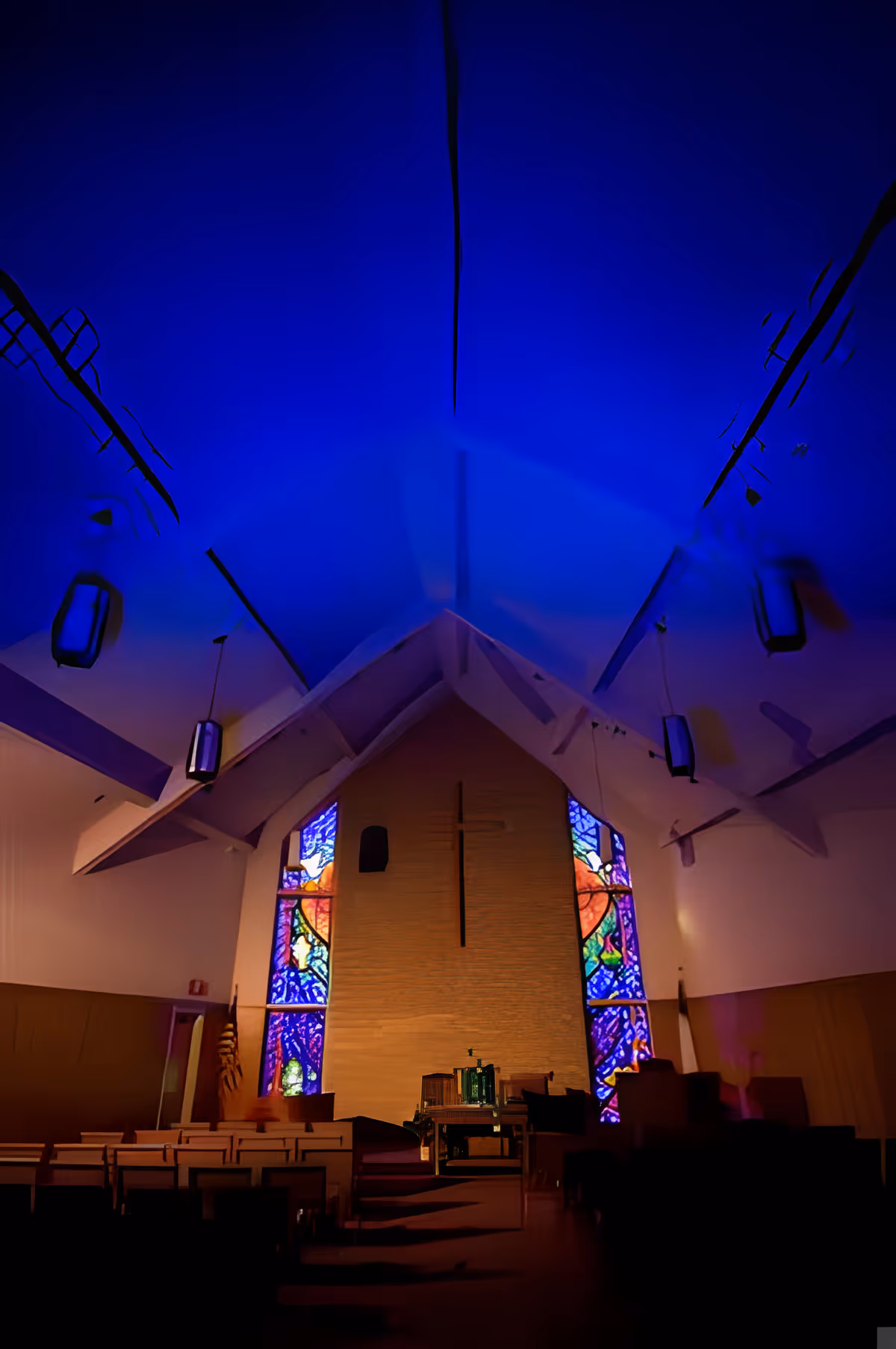 Interior of a chapel with a high vaulted ceiling illuminated in blue light. The front wall features a large cross and colorful stained glass windows on either side. Rows of wooden pews face the altar area, which has chairs and a lectern.
