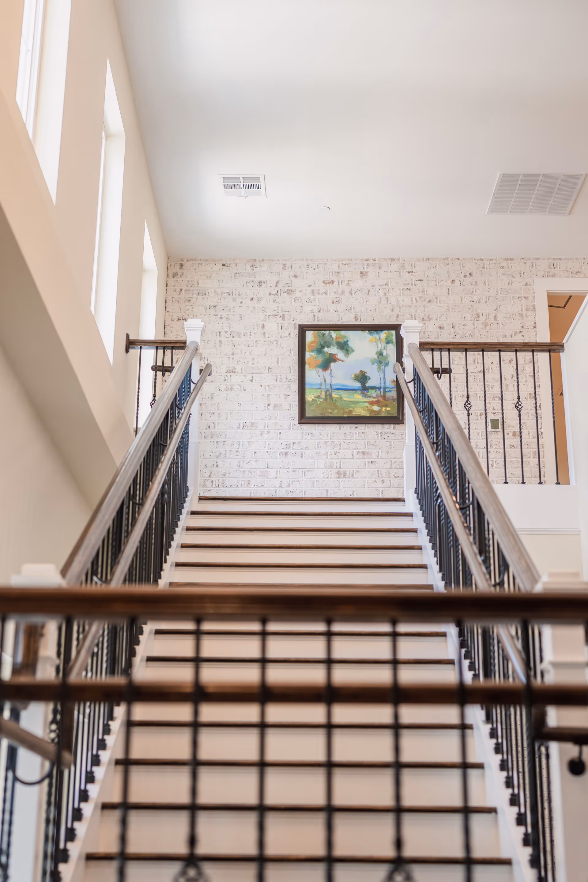A staircase with wooden handrails and black metal balusters leading up to a landing with a white brick wall. A colorful landscape painting is hung on the wall above the stairs. There are three rectangular windows on the left side letting in natural light.