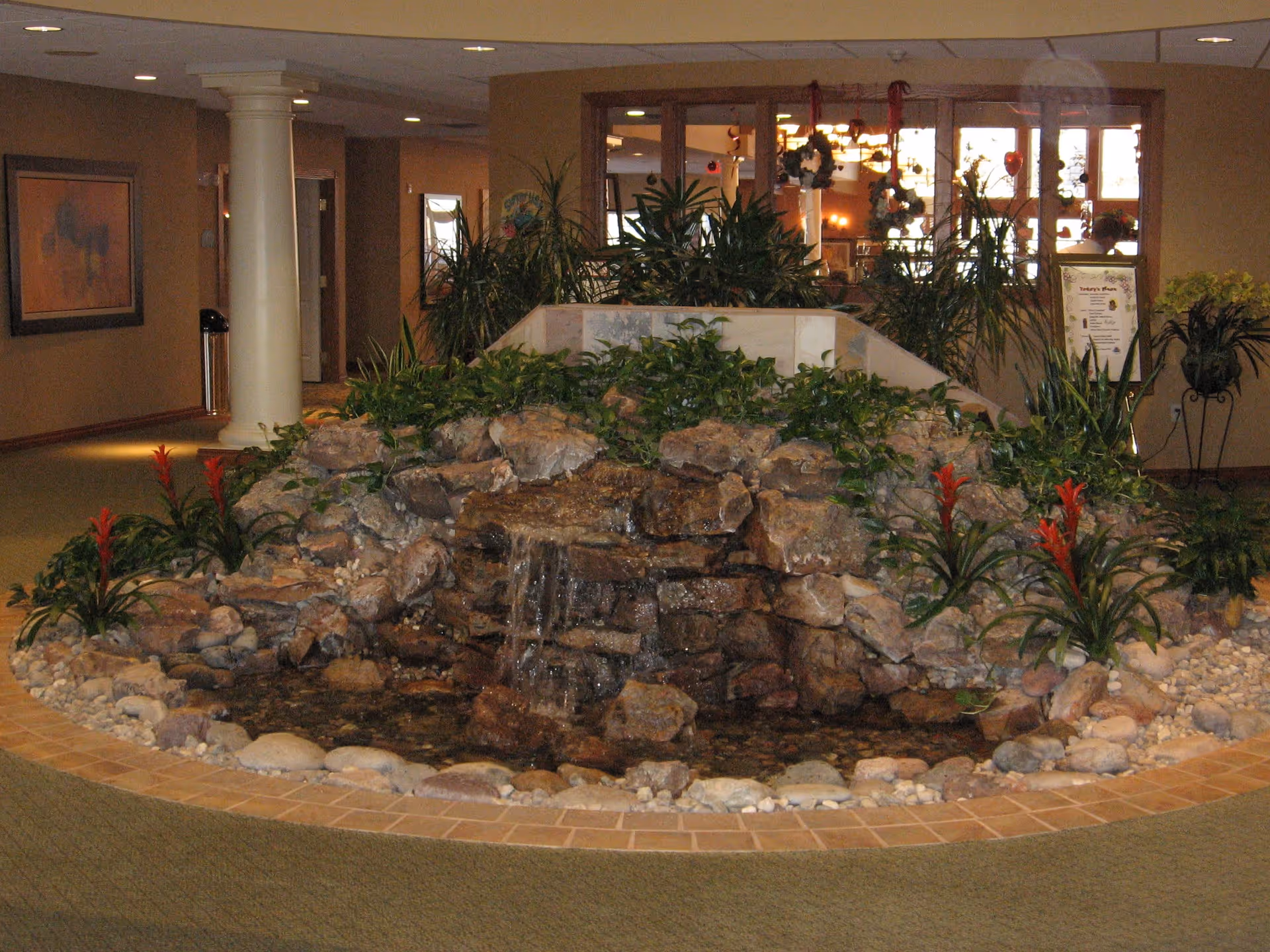 Indoor water feature with rocks, small waterfall, and surrounding plants including red flowers, located in the lobby area of a retirement community with beige walls, carpeted floor, and decorative columns.