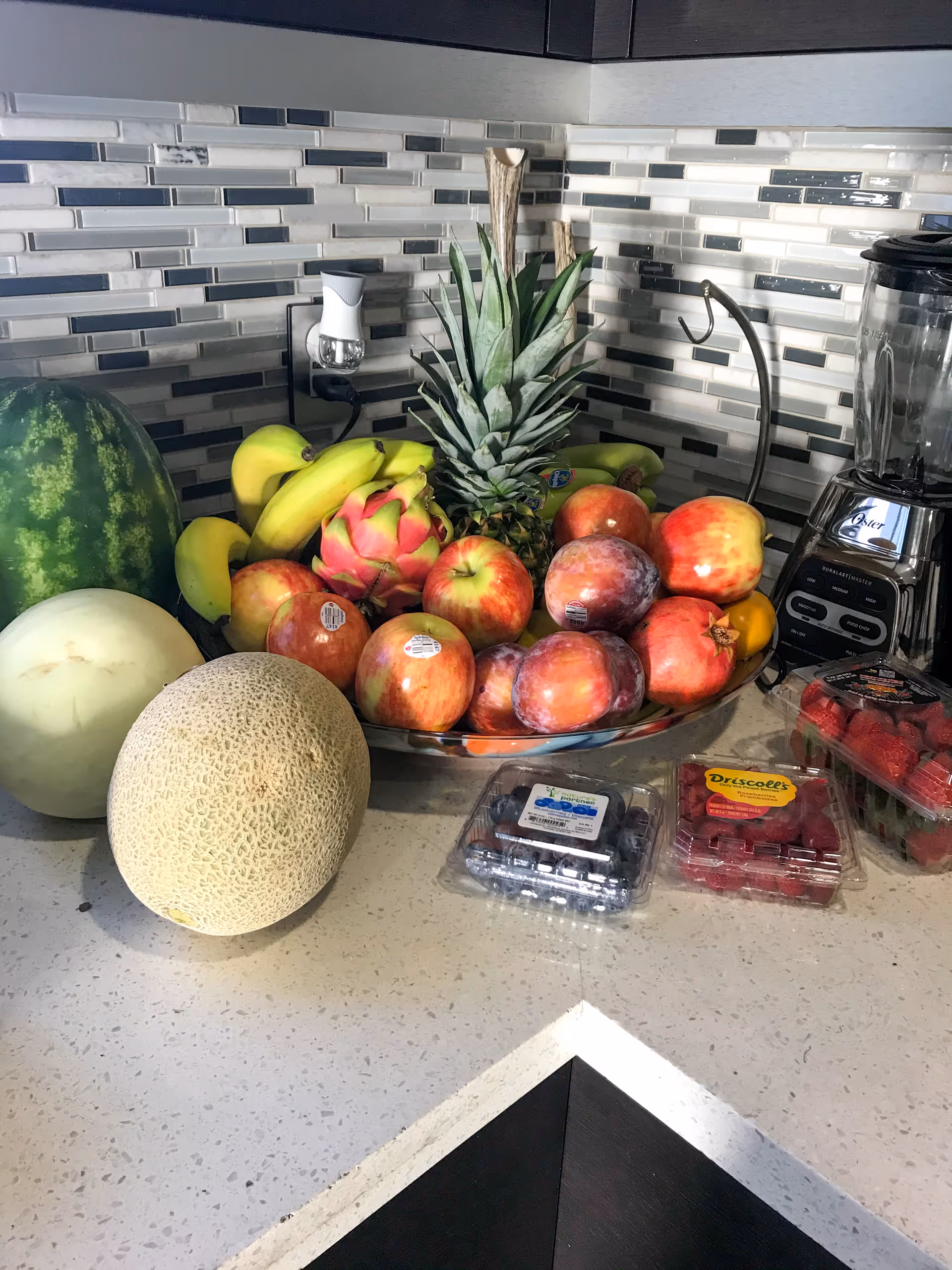 A kitchen countertop with a variety of fresh fruits including a watermelon, cantaloupe, honeydew melon, bananas, pineapple, apples, plums, pomegranate, and containers of blueberries, raspberries, and strawberries. A blender is visible on the right side and a tiled backsplash is in the background.