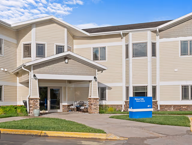 Front entrance of a two-story beige senior living building with a covered entry, outdoor chairs, and a blue sign on the lawn.