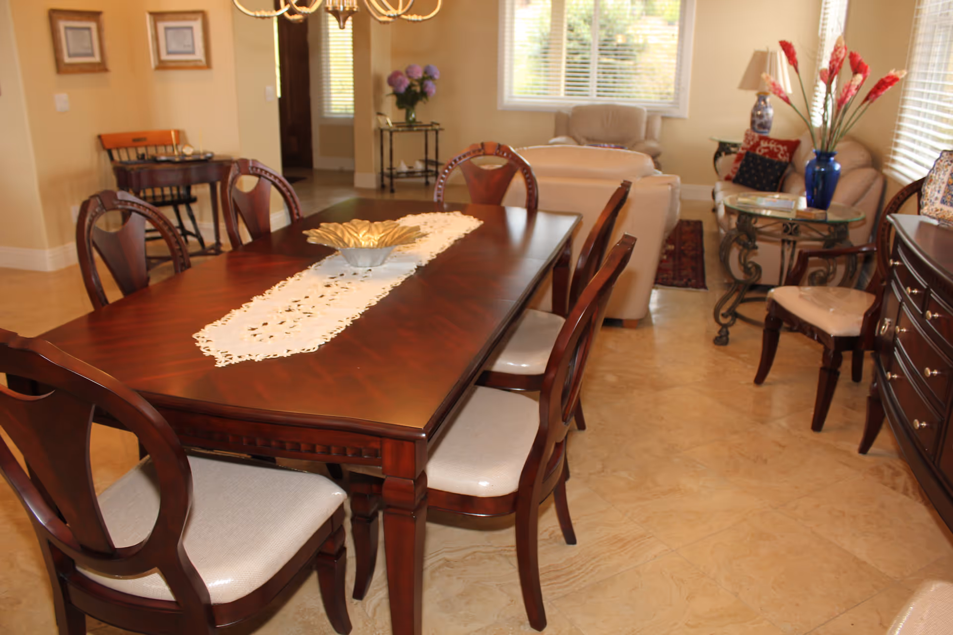 Dining room with a polished wooden table and chairs, a lace runner and centerpiece, opening onto a living area with sofas and side tables.