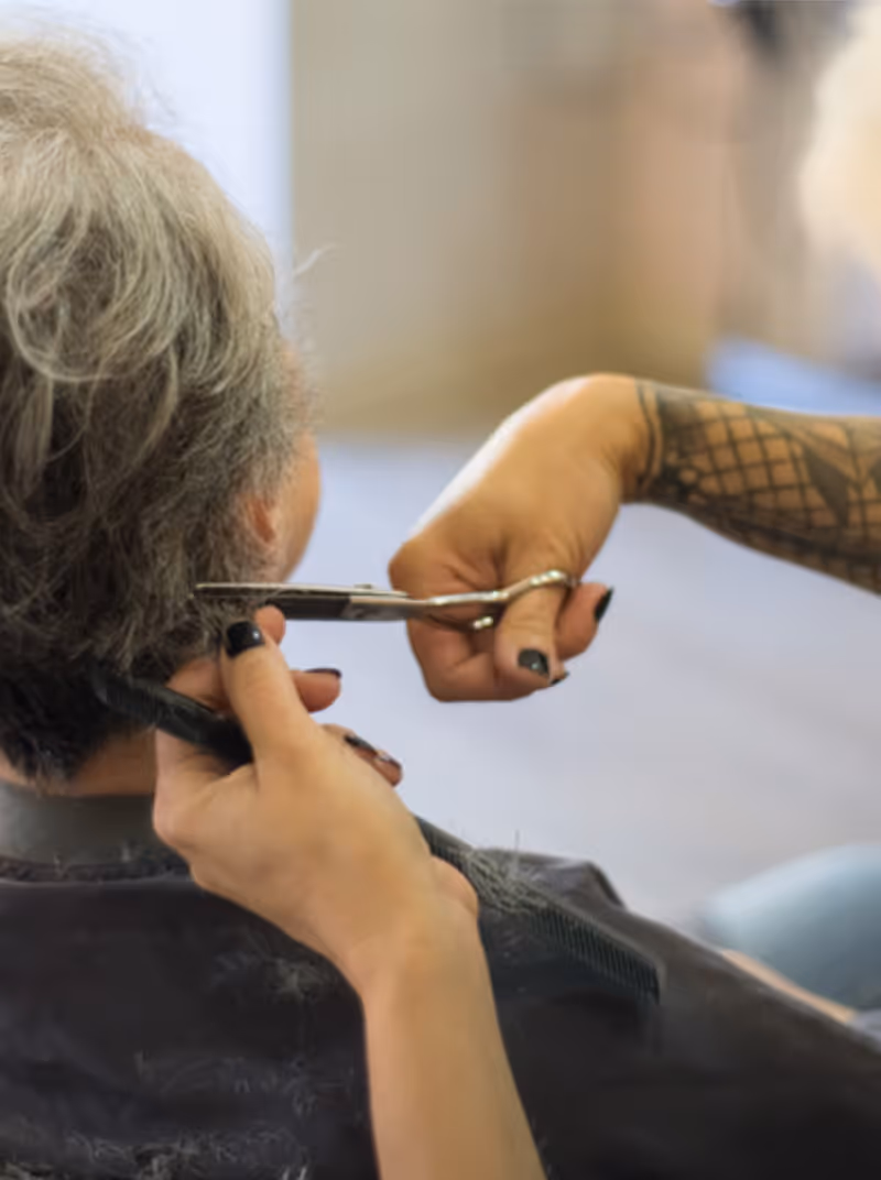 A person with gray hair is getting a haircut. The hairdresser, whose arm with a tattoo is visible, is using scissors and a comb to trim the hair at the back of the person's head.