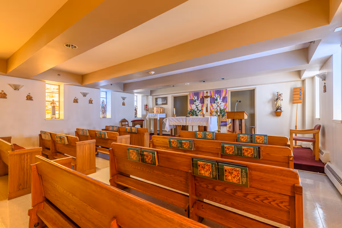 Interior view of a small chapel with wooden pews arranged in rows facing an altar adorned with flowers and religious decorations. The walls have small religious statues and stained glass windows letting in natural light.