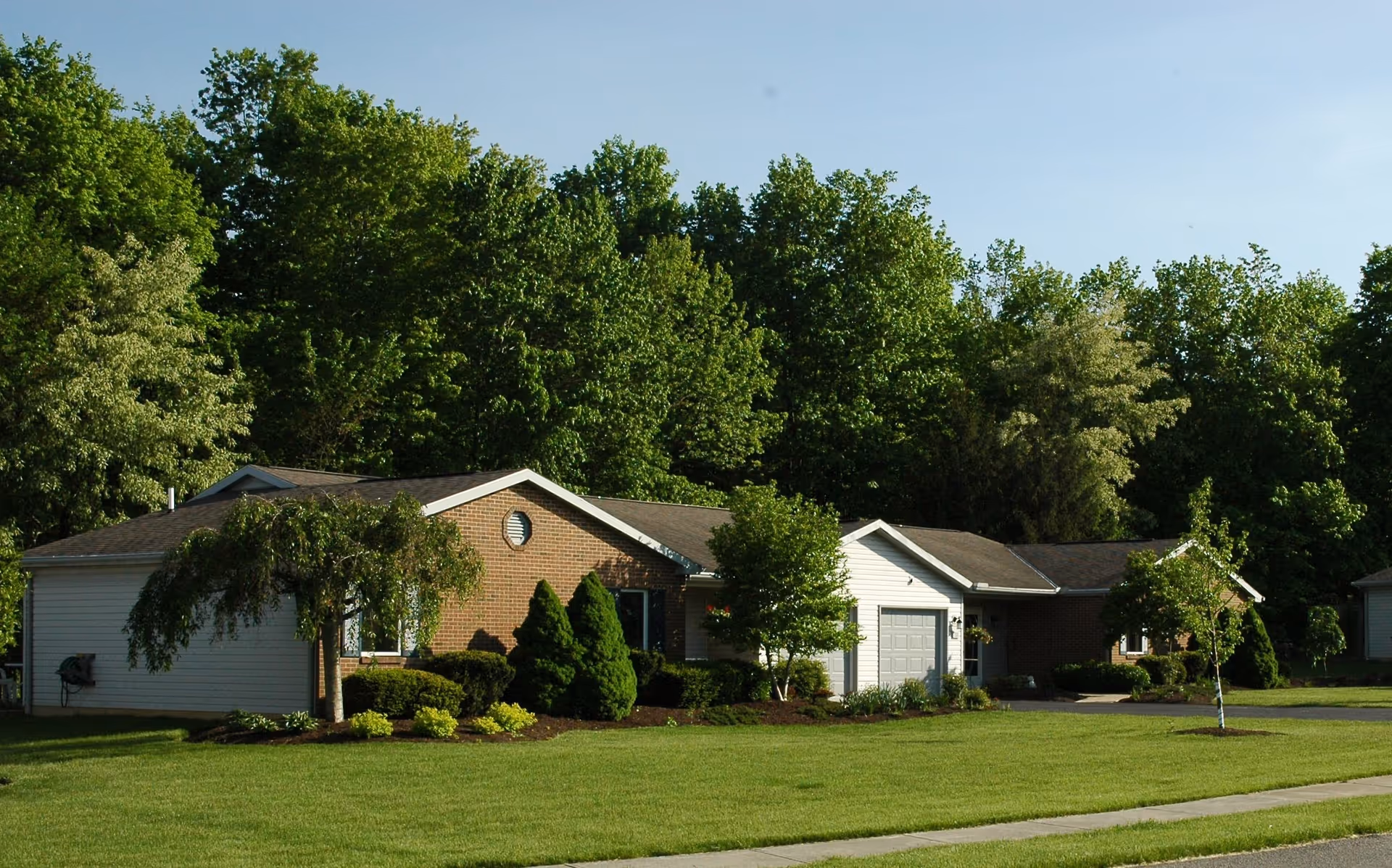 Single-story residential building with a brick and white siding exterior, surrounded by well-maintained green lawn, shrubs, and trees under a clear blue sky.