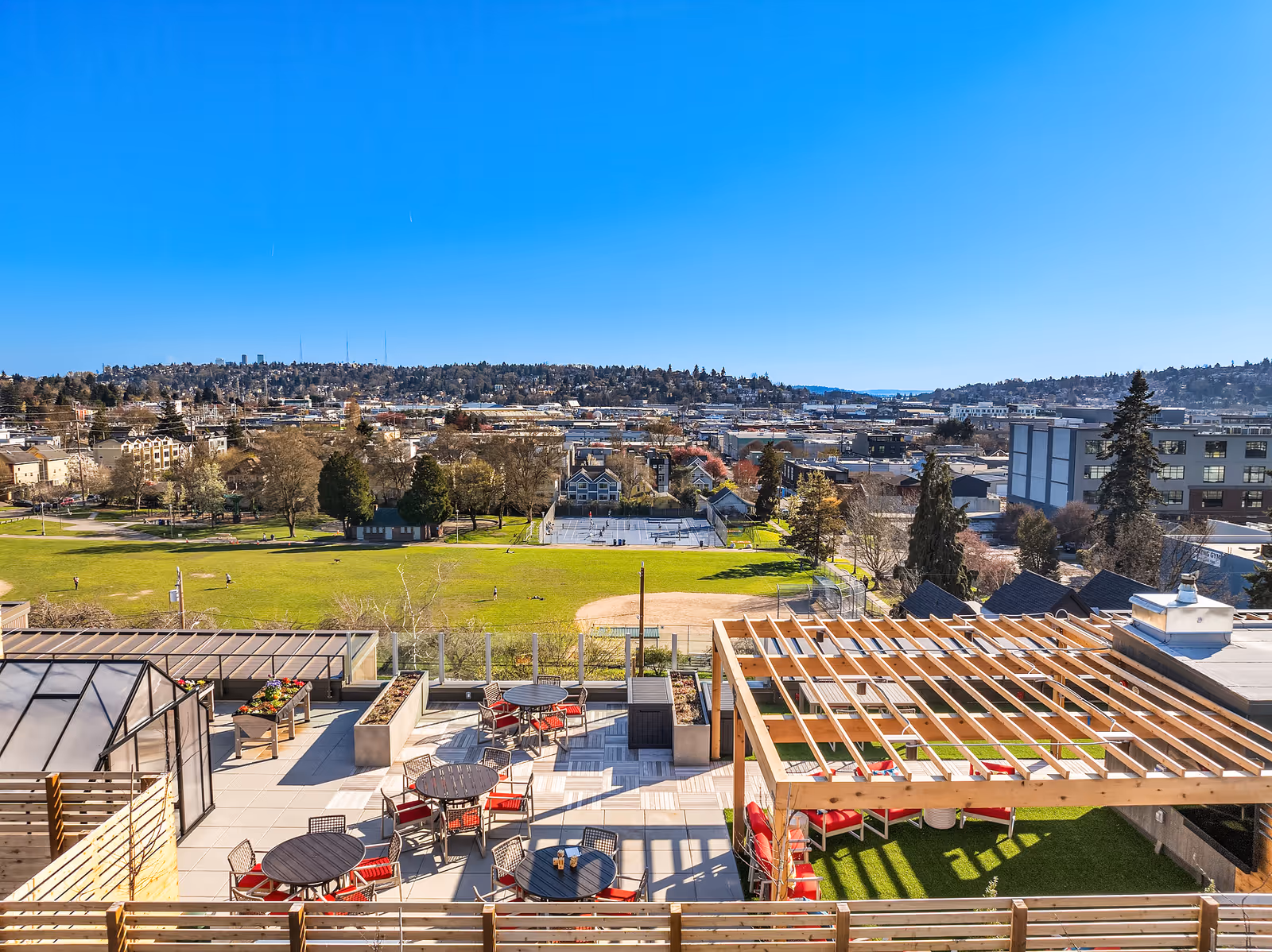 Rooftop patio area with round tables and red cushioned chairs, a wooden pergola, and planter boxes overlooking a large green park with trees, a baseball field, and tennis courts under a clear blue sky.