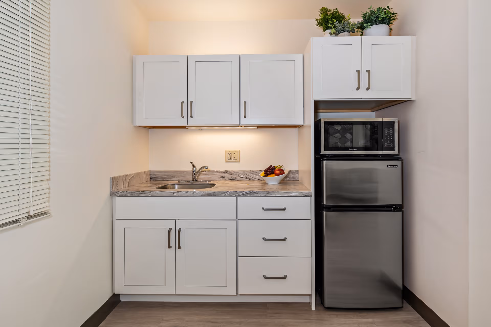 A small kitchen area with white cabinets, a marble countertop, a stainless steel sink, a bowl of fruit, a stainless steel microwave, and a stainless steel mini refrigerator. There are potted plants on top of the upper cabinets and a window with blinds on the left side.