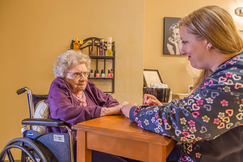 An elderly woman in a wheelchair receiving a manicure from a caregiver in a cozy room with nail polish bottles on a wall shelf and a framed picture on the wall.
