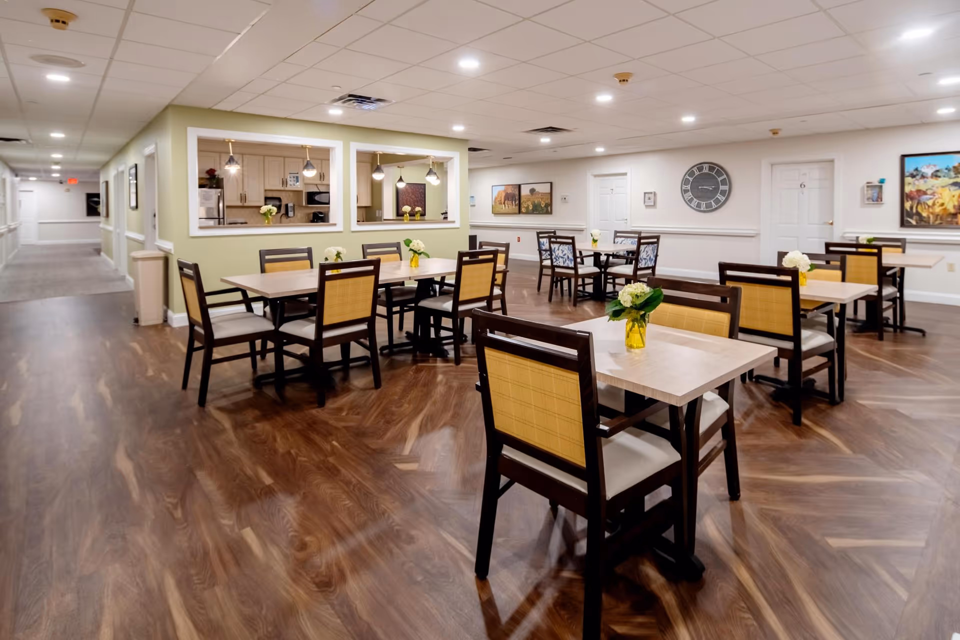 A spacious dining area with several wooden tables and chairs arranged neatly. Each table has a small vase with white flowers. The room has wood-patterned flooring, light-colored walls, and a ceiling with recessed lighting. There are two large windows looking into a kitchen area with pendant lights. The walls are decorated with framed artwork and a large clock. Doors labeled with numbers are visible in the background.