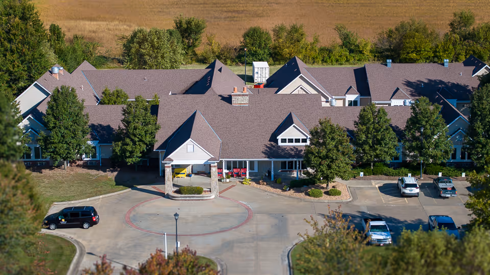 Aerial view of Homestead Assisted & Independent Living of Girard showing a large building with a brown roof surrounded by trees and a parking lot with several cars parked. The building has a covered entrance with a circular driveway in front.