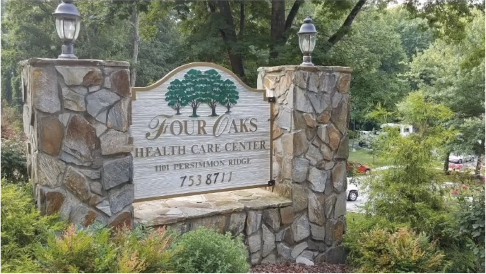 Stone entrance sign reading Four Oaks Health Care Center set among trees and shrubs.