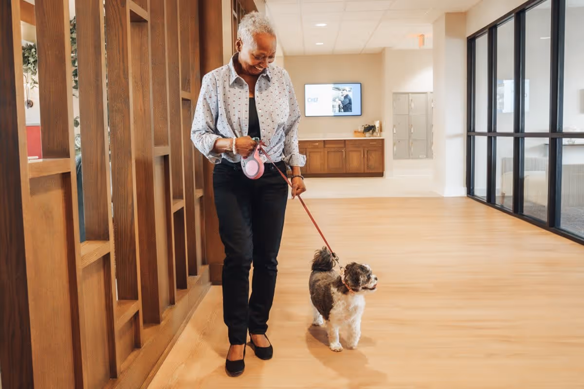 An elderly woman walking a small dog on a leash inside a spacious, well-lit hallway with wooden flooring and modern decor.