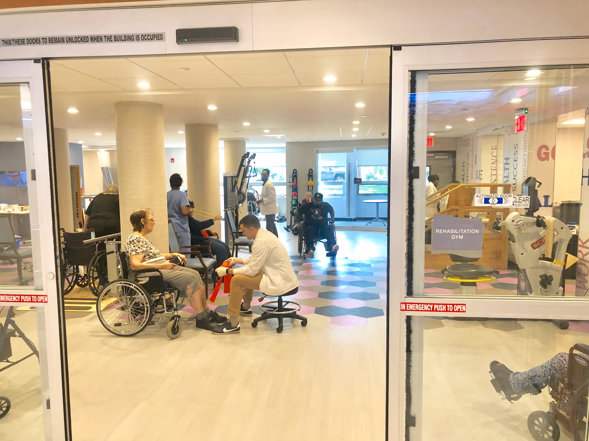 View through glass doors into a rehabilitation gym where elderly individuals, some in wheelchairs, are engaged in physical therapy activities with staff assistance. The room has exercise equipment, bright lighting, and a patterned floor.