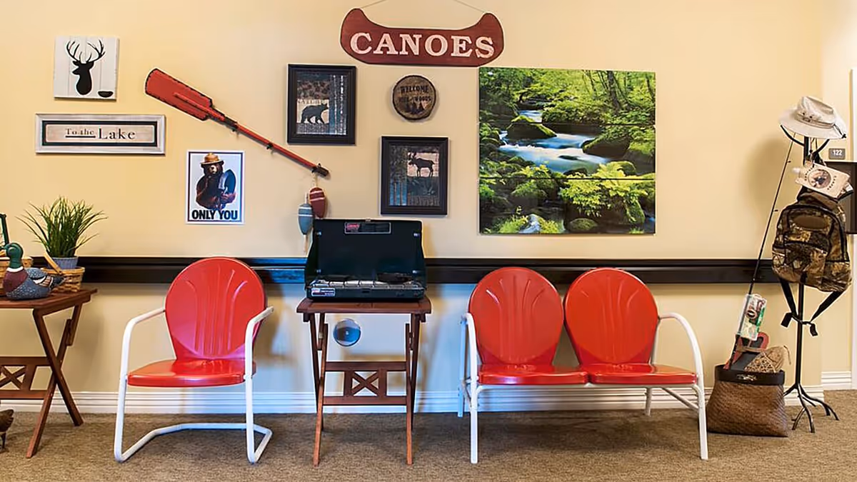 Indoor seating area with two red metal chairs and one red metal bench against a beige wall. The wall is decorated with various nature-themed pictures, a red canoe paddle, and a wooden sign that says 'CANOEES'. A small table with a portable stove is placed between the chairs. To the right, there is a coat rack holding hats, a backpack, and other outdoor gear. A small table with a plant and decorative items is on the left side.