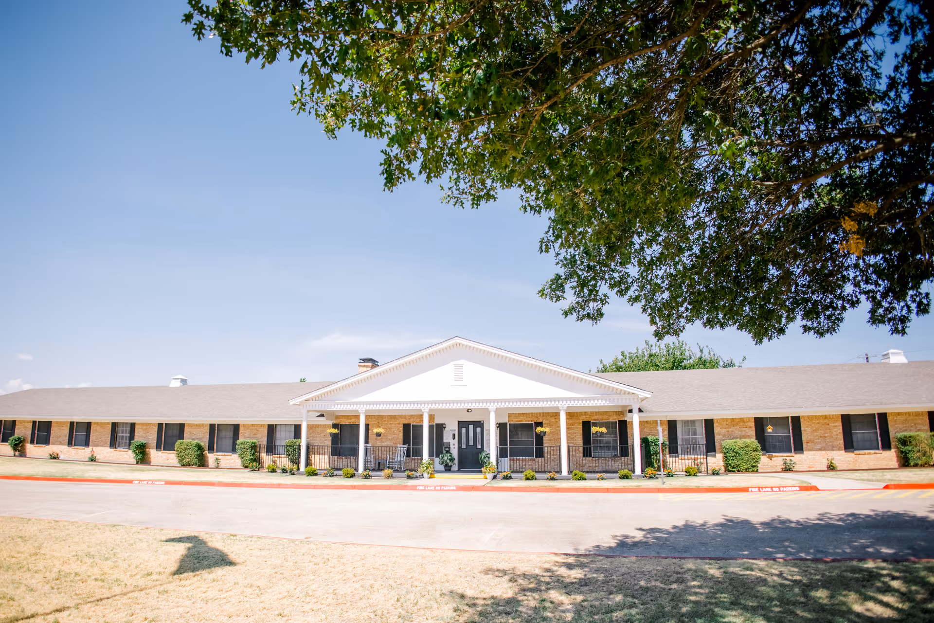Front exterior view of a single-story nursing and rehabilitation facility building with a covered entrance, brick walls, multiple windows with black shutters, and a large tree providing shade in the foreground.