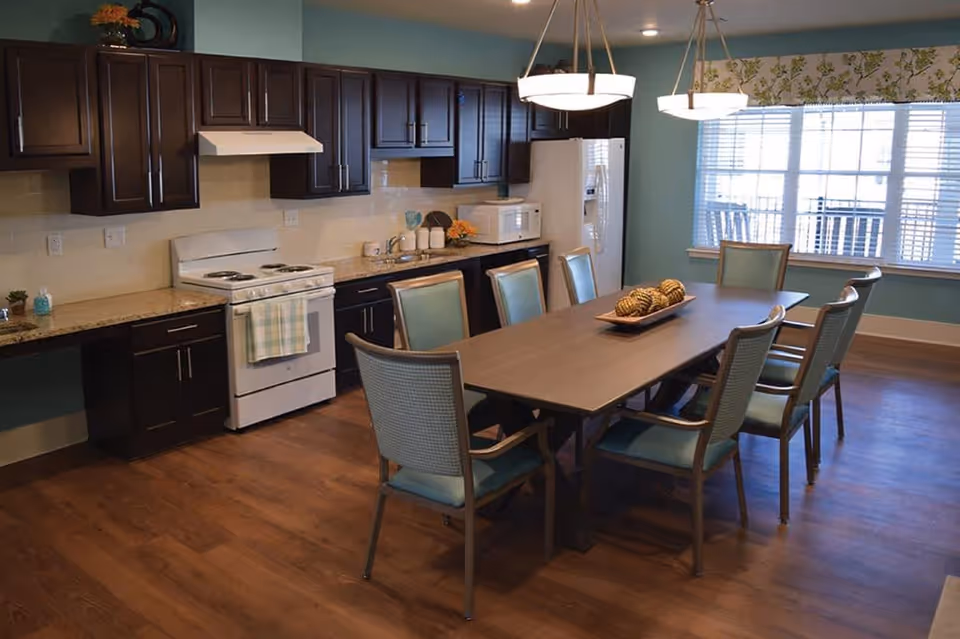 A spacious kitchen and dining area with dark wood cabinets, a white stove and refrigerator, and a long dining table surrounded by eight chairs with light blue cushions. The room has wooden flooring, two hanging light fixtures above the table, and a large window with blinds and a floral valance.