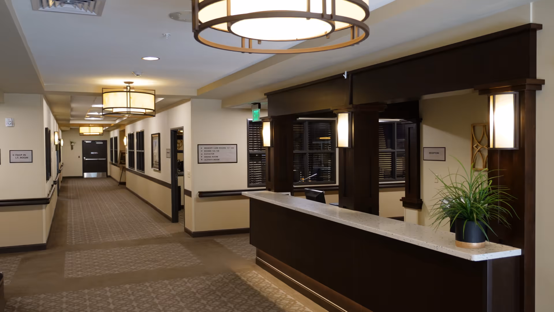 Interior view of a senior living facility hallway with beige walls and carpeted floor. On the right side, there is a dark wood reception desk with a white countertop and a potted plant. The hallway is well-lit with ceiling lights and wall sconces. Several doors and framed signs are visible along the hallway.