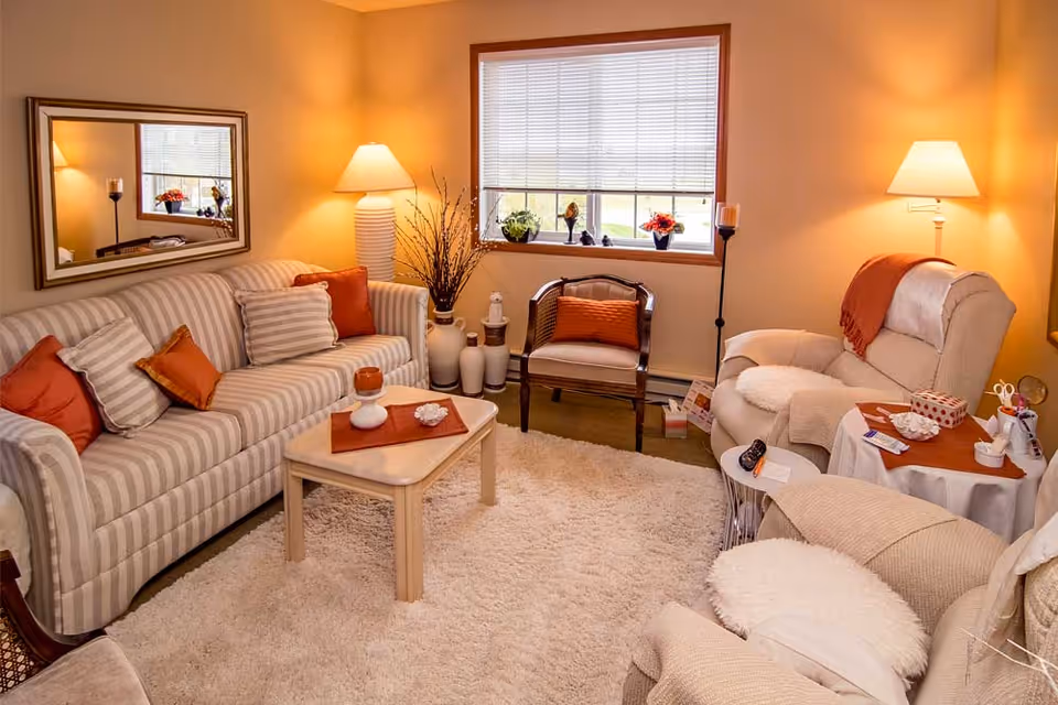 A cozy living room with a striped sofa adorned with orange and beige pillows, a wooden chair with an orange cushion, and two beige armchairs. The room features a soft beige carpet, a small wooden coffee table with decorative items, a large window with blinds, two floor lamps, and various vases and decor pieces. A large mirror hangs on the wall above the sofa.
