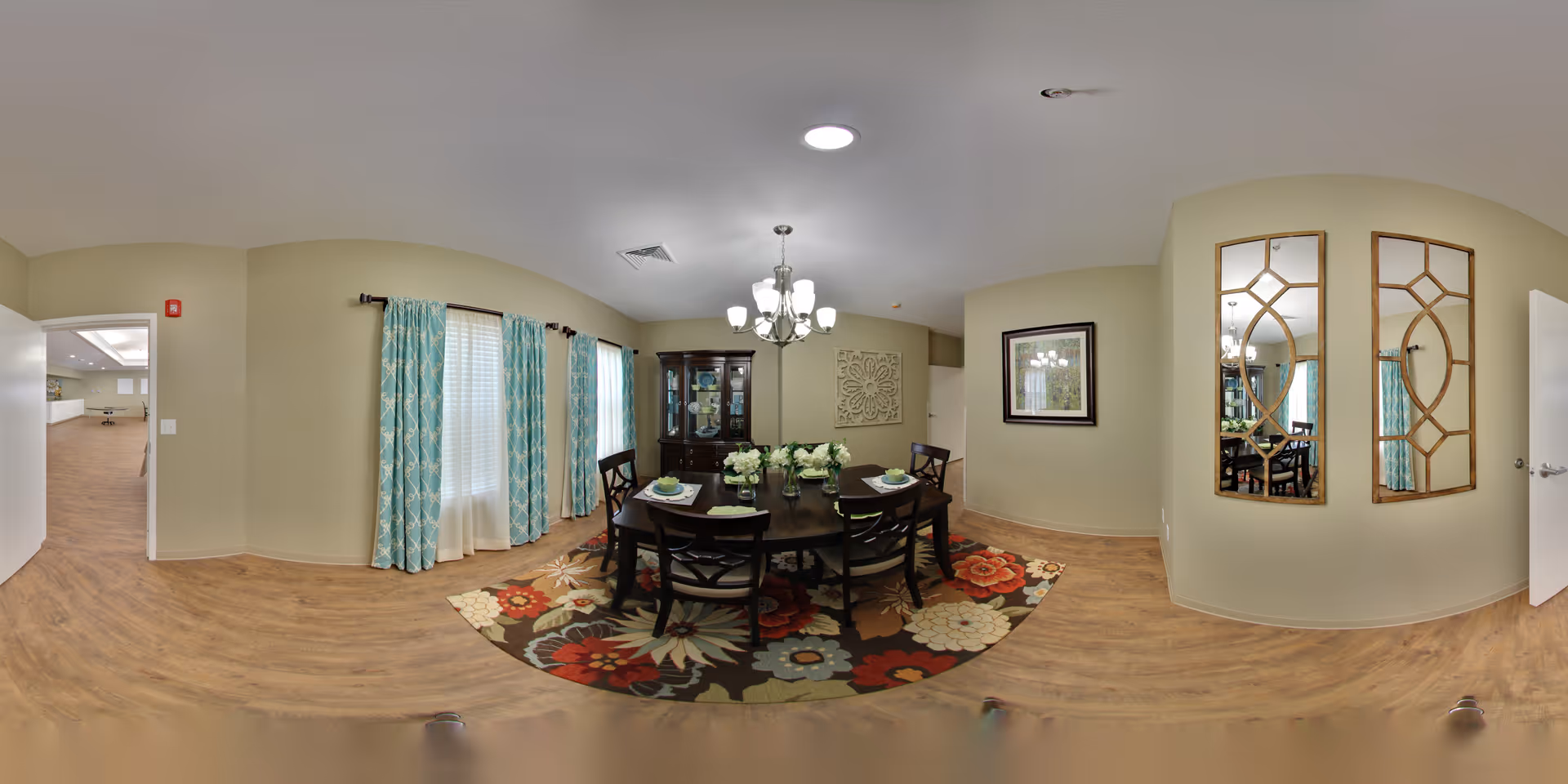 A dining room with a dark wooden table set for four, surrounded by matching chairs. The table is decorated with a floral centerpiece and place settings. The room features light-colored walls, wood flooring, a colorful floral area rug under the table, and two large windows with blue patterned curtains. A chandelier hangs above the table, and a dark wooden china cabinet is positioned against the far wall. Two decorative mirrors with geometric wooden frames are mounted on one wall, and a framed picture hangs on another. An open door leads to an adjacent room with more wood flooring.