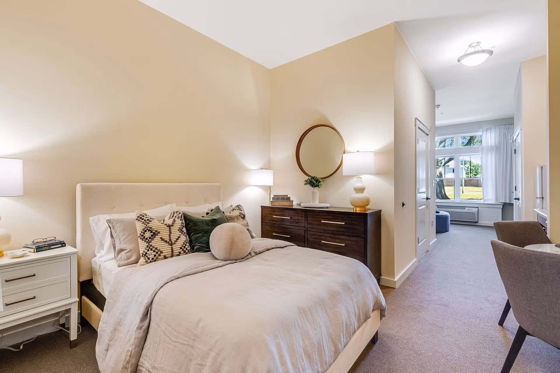 A cozy bedroom in a senior living facility with a neatly made bed featuring multiple pillows, a white nightstand with a lamp and books, a dark wooden dresser with a round mirror and lamp, and a window with white curtains letting in natural light.