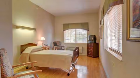 A furnished hospital-style bedroom with bed, chair, dresser, and windows.