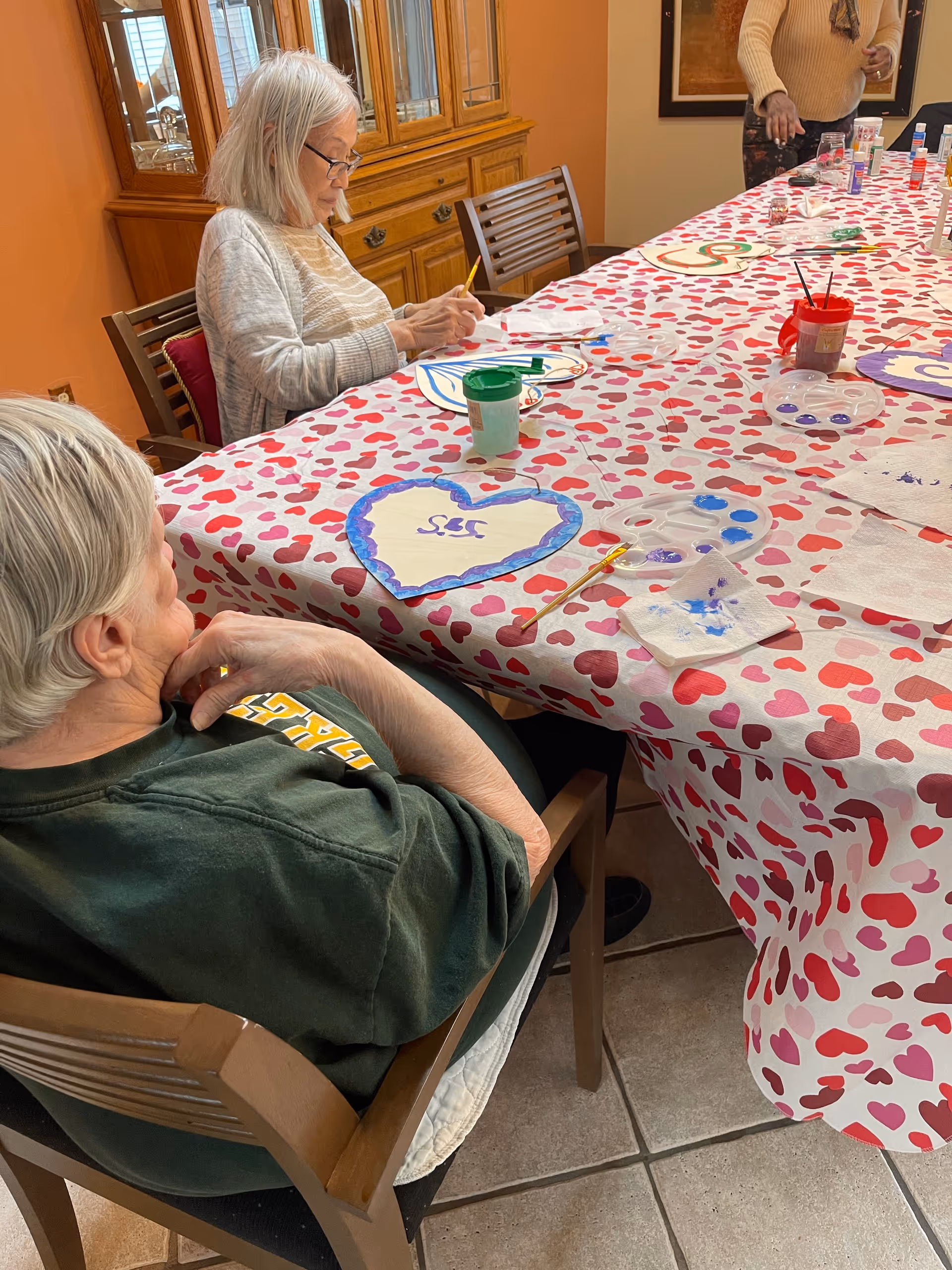 Elderly residents seated at a long table with a heart-patterned tablecloth painting heart-shaped crafts in a communal dining/activity room.
