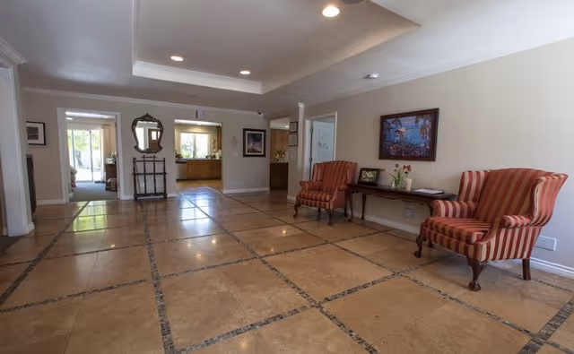 A spacious interior common area with tiled flooring and recessed lighting. The room features two red and beige striped armchairs placed against a wall with a framed painting above a wooden console table holding a vase with flowers and some papers. There is a large mirror and a decorative stand near the entrance to another room with windows letting in natural light.