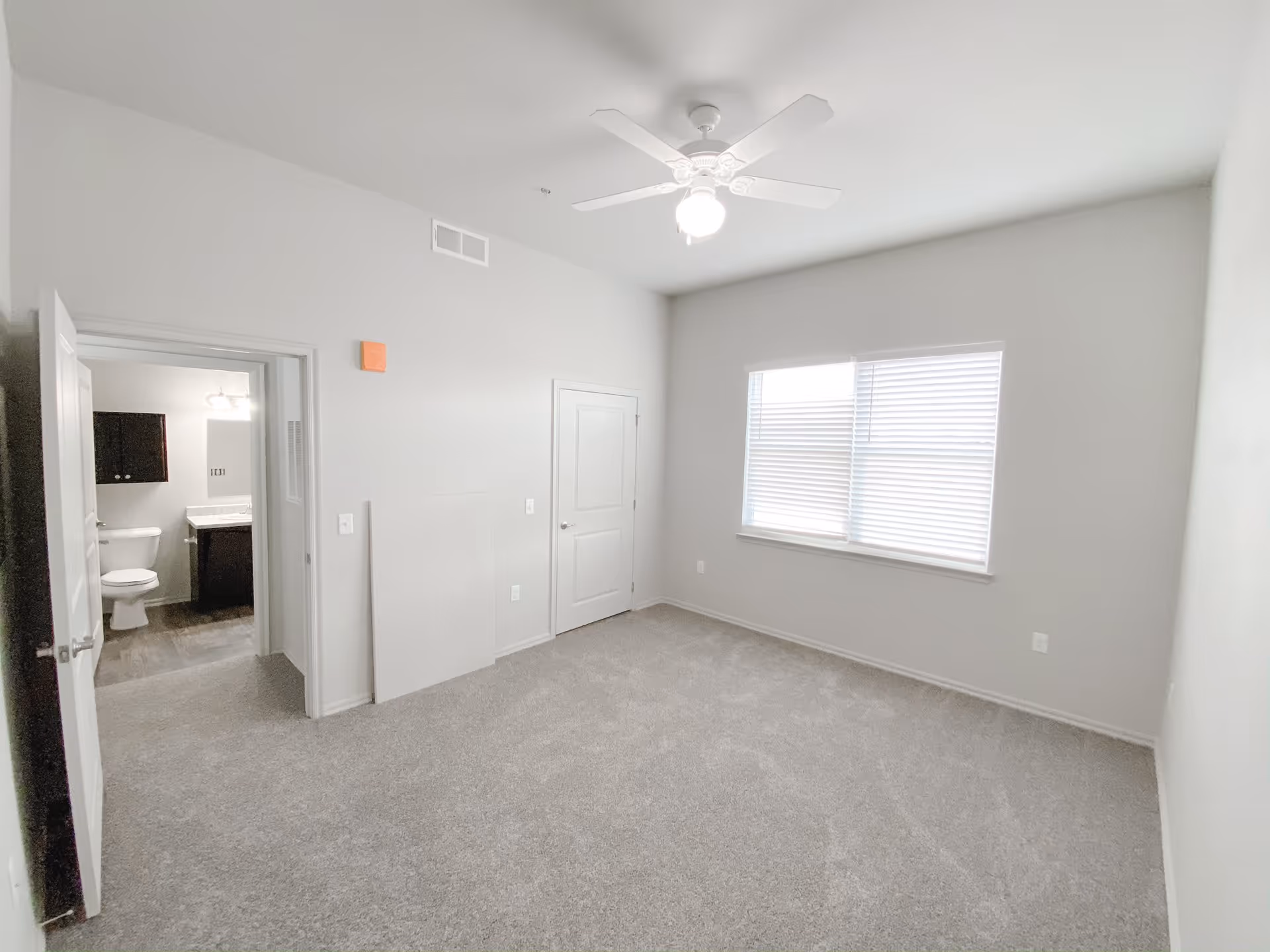 Empty bedroom with light gray carpet, white walls, a ceiling fan with light, a window with blinds, and an open door leading to a bathroom with a toilet and sink.