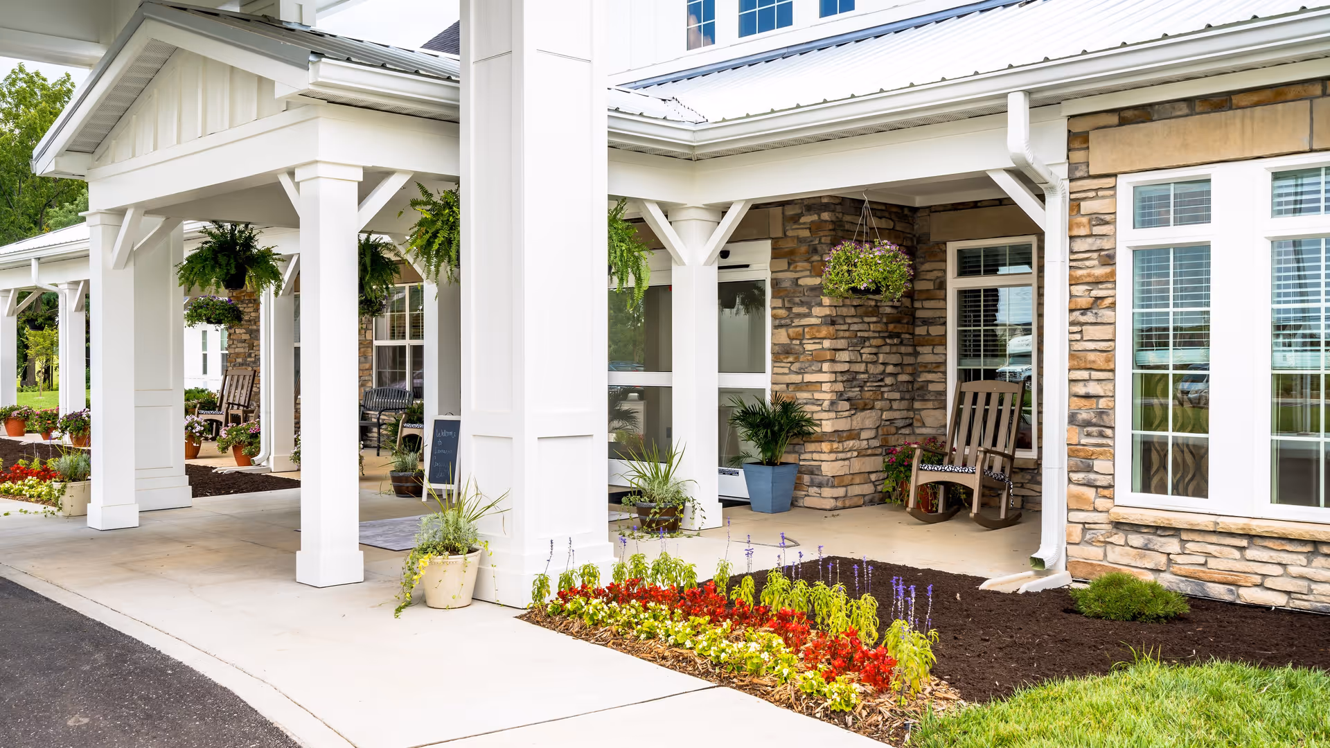 Entrance area of Demaree Crossing facility featuring a covered porch with white pillars, hanging flower baskets, potted plants, a rocking chair, and a flower bed with red, yellow, and purple flowers next to a stone and siding exterior wall.